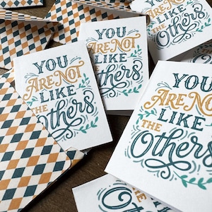 May include: Greeting cards with the message "You Are Not Like the Others" in teal and gold script. The cards are white with decorative floral accents. Envelopes with a diamond pattern in teal, gold, and white are also visible.