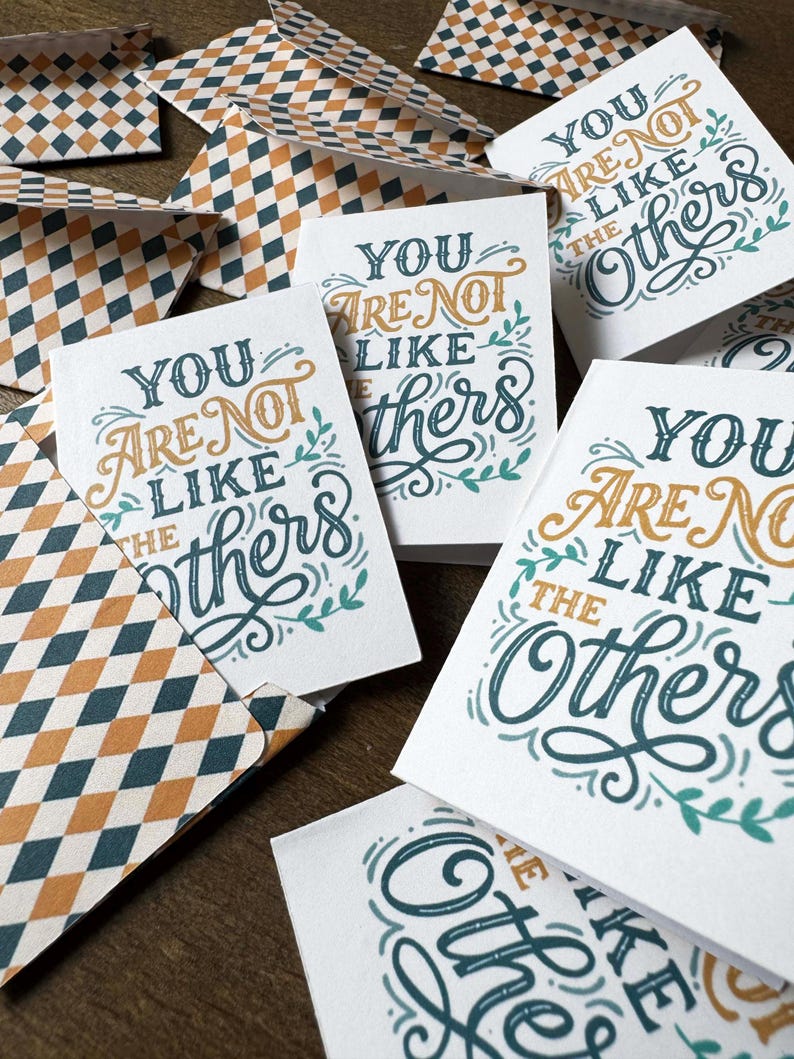 May include: Greeting cards with the message "You Are Not Like the Others" in teal and gold script. The cards are white with decorative floral accents. Envelopes with a diamond pattern in teal, gold, and white are also visible.