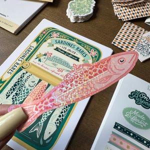 May include: A pink fish-shaped sticker with iridescent glitter is held over a Sardines Rares label. The label features a fish illustration and text. Other stickers and stationery items are scattered around.