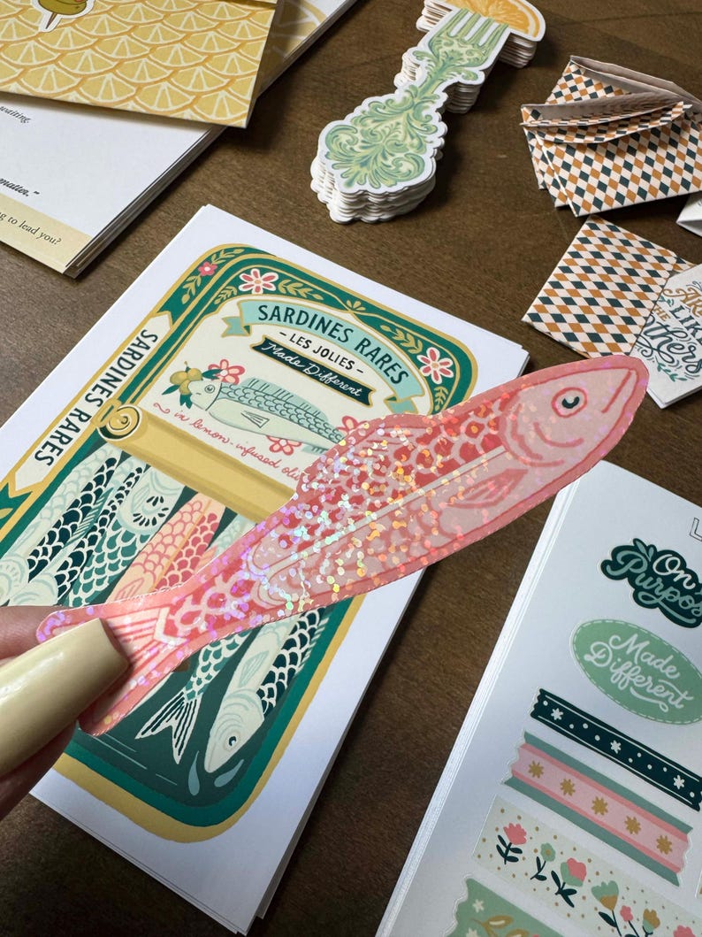 May include: A pink fish-shaped sticker with iridescent glitter is held over a Sardines Rares label. The label features a fish illustration and text. Other stickers and stationery items are scattered around.