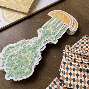 May include: A stack of stickers featuring a decorative fork design with a lemon slice on top. The fork has a green and blue ornate pattern. Other stationery items include a lemon-themed envelope and diamond-patterned envelopes. The text "Majestic Crumbs" is visible.