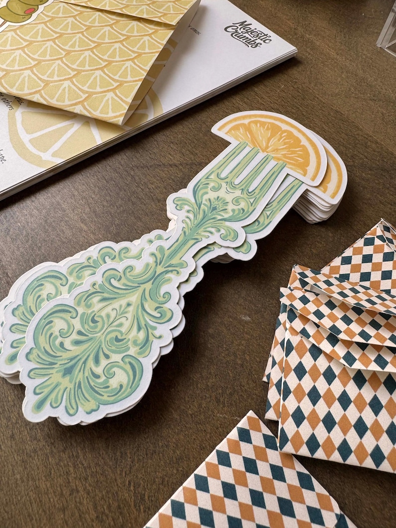 May include: A stack of stickers featuring a decorative fork design with a lemon slice on top. The fork has a green and blue ornate pattern. Other stationery items include a lemon-themed envelope and diamond-patterned envelopes. The text "Majestic Crumbs" is visible.