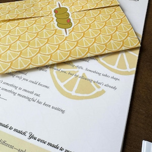 May include: A yellow envelope with a lemon slice pattern and an olive sticker rests on a white paper with text. The envelope is labeled "May 2026" and has the words "Majestic Crumbs" printed on the paper.