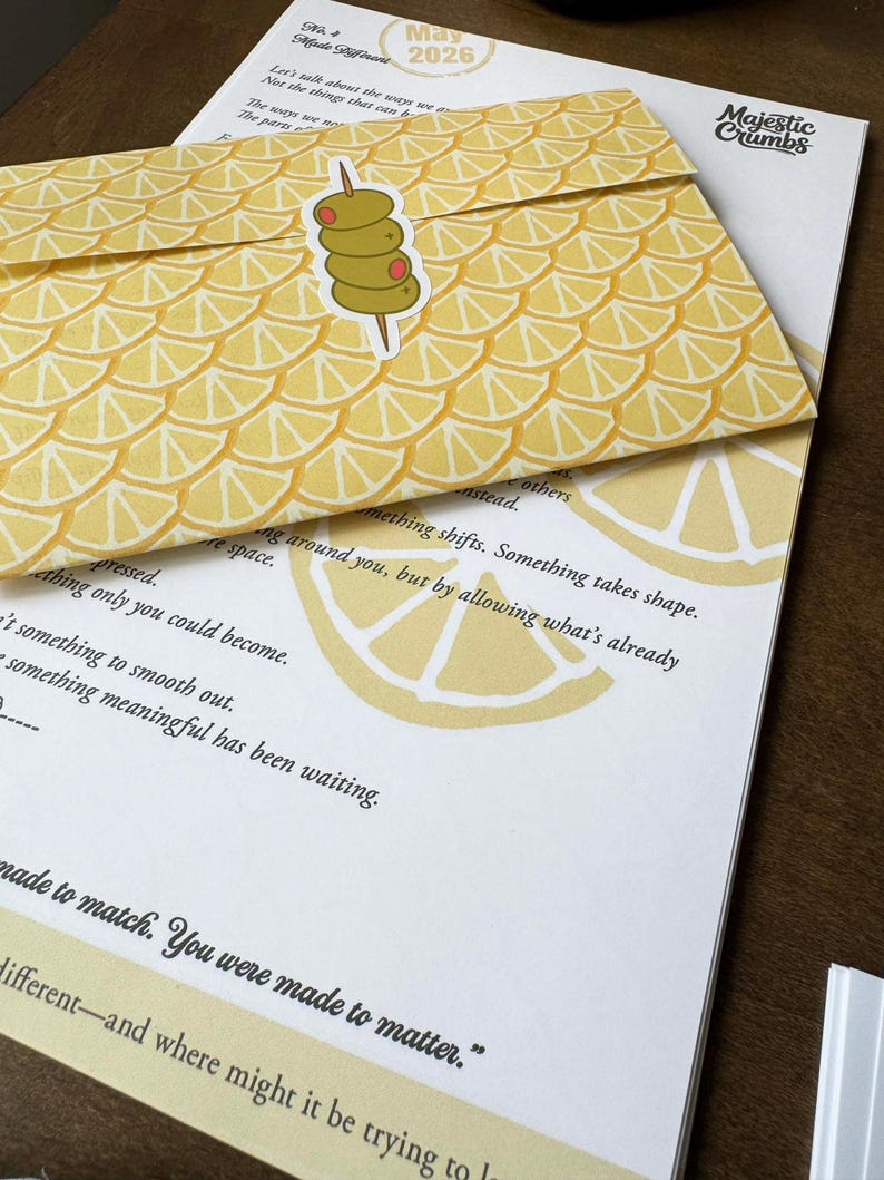 May include: A yellow envelope with a lemon slice pattern and an olive sticker rests on a white paper with text. The envelope is labeled "May 2026" and has the words "Majestic Crumbs" printed on the paper.