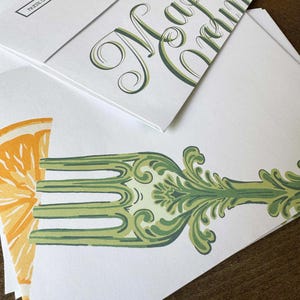 May include: White stationery with green and orange illustrations. One envelope features the words "Majestic Crumbs" in elegant script. Another shows a fork design with an orange slice. The stationery is stacked on a wooden surface.