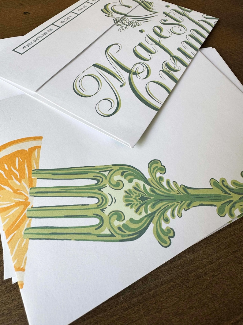 May include: White stationery with green and orange illustrations. One envelope features the words "Majestic Crumbs" in elegant script. Another shows a fork design with an orange slice. The stationery is stacked on a wooden surface.