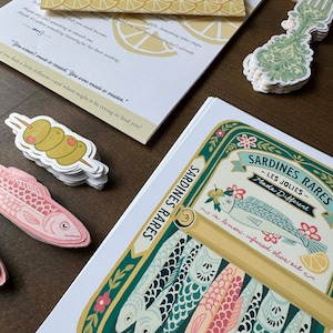 May include: An assortment of paper goods and stickers, including a sardine illustration, olive skewers, and a decorative fork. The sardine illustration features colorful fish in a can with text that reads "Sardines Rares - Les Jolies - Made Different".