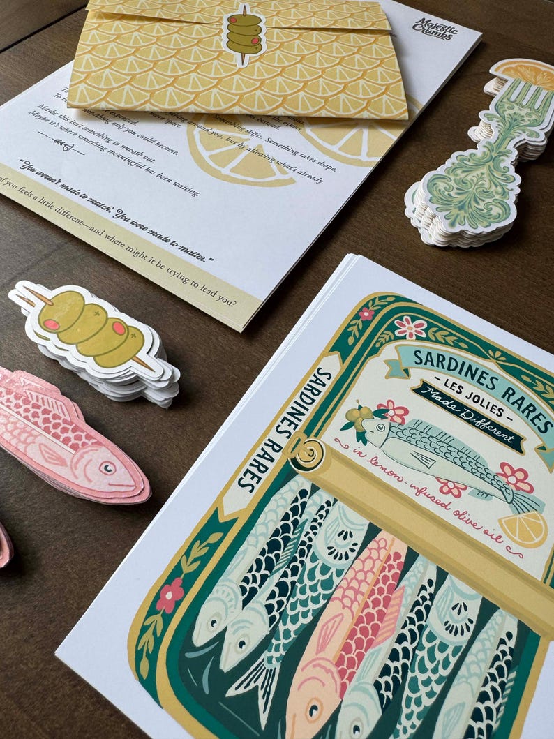 May include: An assortment of paper goods and stickers, including a sardine illustration, olive skewers, and a decorative fork. The sardine illustration features colorful fish in a can with text that reads "Sardines Rares - Les Jolies - Made Different".