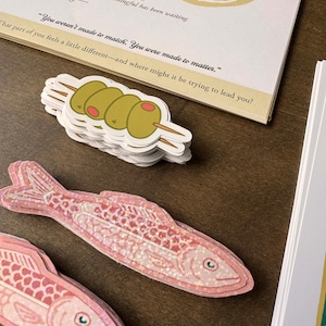 May include: A stack of olive stickers on a wooden surface. The stickers feature three green olives on a wooden skewer. A pink fish sticker with a glittery pattern is also visible, along with a printed card with text.