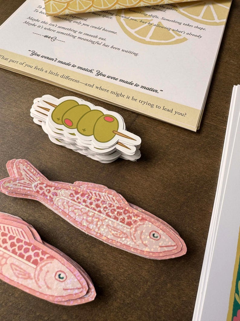 May include: A stack of olive stickers on a wooden surface. The stickers feature three green olives on a wooden skewer. A pink fish sticker with a glittery pattern is also visible, along with a printed card with text.