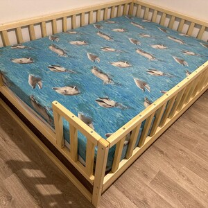 May include: A wooden toddler bed with a blue and white dolphin print sheet. The bed has a safety rail on all sides.