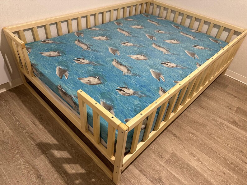 May include: A wooden toddler bed with a blue and white dolphin print sheet. The bed has a safety rail on all sides.