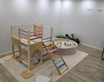 May include: A wooden indoor climbing structure featuring a multi-colored ladder, a climbing net, and circular cutouts. Beside it sits a round ball pit filled with white and grey balls, a green toy tractor, and a white wooden ride-on toy cart.