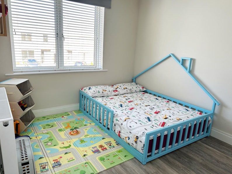 May include: A light blue toddler bed with a house-shaped headboard and a white and blue patterned bedding. The bed has a safety rail on one side and is on a light gray floor. A play mat with a road and animal designs is in front of the bed.