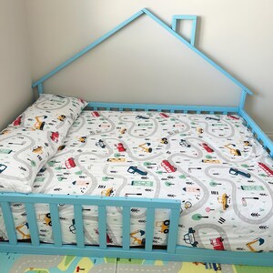 May include: A light blue wooden toddler bed shaped like a house with a white and gray patterned sheet and a matching pillow. The sheet features a design of winding roads with small cars and trucks.