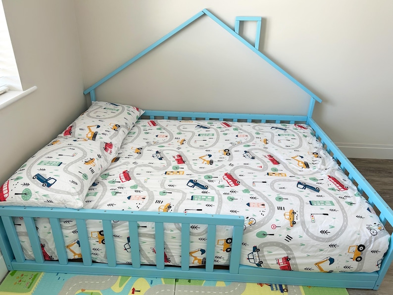 May include: A light blue wooden toddler bed shaped like a house with a white and gray patterned sheet and a matching pillow. The sheet features a design of winding roads with small cars and trucks.