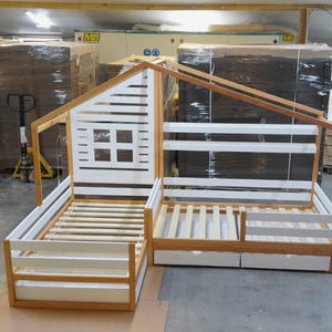 May include: A wooden children's bed shaped like a house with a white painted front and a slatted mattress. The bed has a built-in shelf and a drawer underneath.