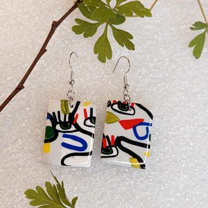 Peut inclure: Une paire de boucles d'oreilles rectangulaires pendantes avec une base blanche ornée de lignes noires abstraites, incluant des formes d'yeux stylisées et des accents géométriques aux couleurs primaires rouge, bleu, jaune et vert, sur crochets argentés.