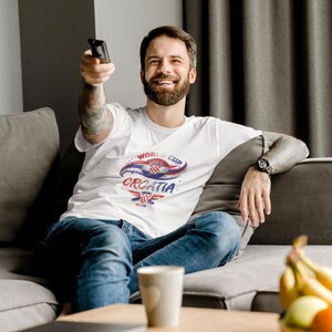 May include: A white short-sleeved t-shirt featuring a graphic design with the text World Cup Croatia, a red and white checkered emblem, and blue and red swooping accents. The shirt is worn with blue denim jeans and a black wristwatch on the wrist.