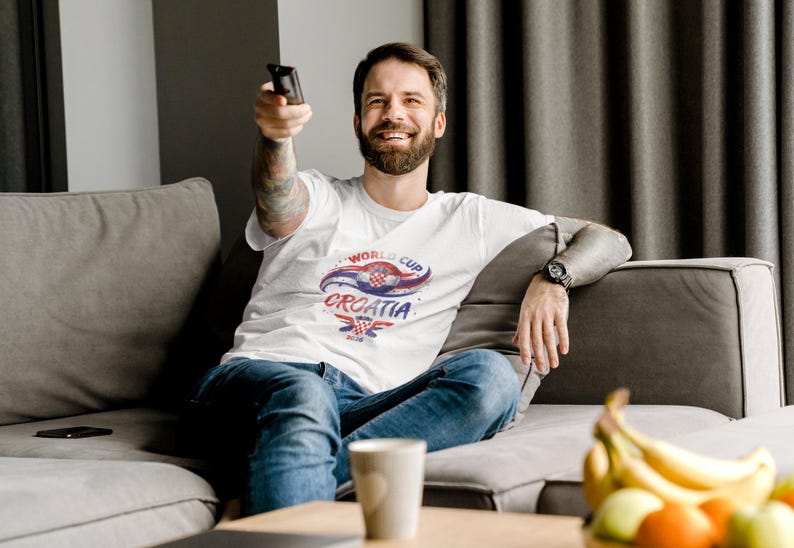 May include: A white short-sleeved t-shirt featuring a graphic design with the text World Cup Croatia, a red and white checkered emblem, and blue and red swooping accents. The shirt is worn with blue denim jeans and a black wristwatch on the wrist.