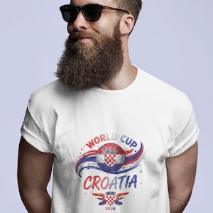 May include: A white short-sleeved t-shirt featuring a graphic print of a soccer ball with a red and white checkered pattern, blue and red swooshes, and the text "WORLD CUP CROATIA 2026" with a small emblem of a checkered shield and wings.