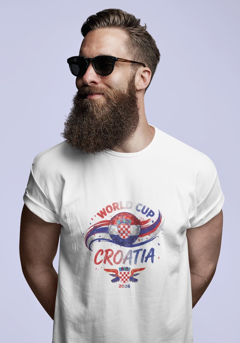 May include: A white short-sleeved t-shirt featuring a graphic print of a soccer ball with a red and white checkered pattern, blue and red swooshes, and the text "WORLD CUP CROATIA 2026" with a small emblem of a checkered shield and wings.