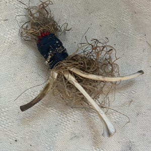 May include: A small, handmade doll made from natural materials. The doll has a blue and white fabric body, red beads, and long, thin, white bone-like legs. The doll is surrounded by dried, brown, stringy material.