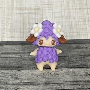 May include: A handcrafted figurine of a sheep with a purple wool coat, brown ears, and large, expressive eyes. The sheep is adorned with white flower accents and stands on a cream-colored base. The figurine is likely made of polymer clay.