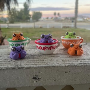 May include: A collection of colorful frog figurines, each with large eyes and a smiling expression. The frogs are in various colors, including orange, green, and purple. Some are nestled in decorative teacups with floral patterns. The scene is set outdoors.
