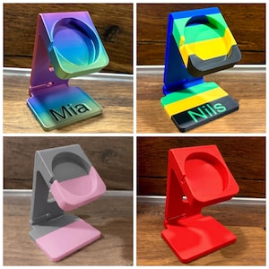 Puede incluir: Cuatro soportes de carga en forma de L para relojes inteligentes. Uno es de color arcoíris con el nombre Mia, uno es azul, verde, amarillo y negro con el nombre Nils, uno es gris y rosa, y uno es rojo sólido. Cada soporte tiene un hueco circular.