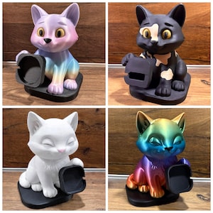 May include: Four cat-shaped phone stands on black bases. One is pastel gradient, one is black with white patches, one is solid white, and one is a metallic rainbow gradient. Each cat figure holds a black square holder for a mobile device.