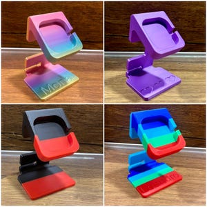 Könnte beinhalten: Eine Sammlung von 3D-gedruckten Telefonständern in verschiedenen Farben. Ein Ständer ist ein Farbverlauf aus Rosa, Gelb und Blau mit dem Aufdruck "Mona". Ein anderer ist einfarbig lila mit Herzformen. Die anderen sind rot, schwarz und blau.