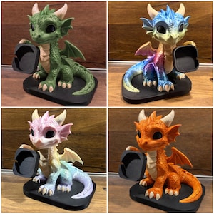 Pode incluir: Uma coleção de quatro figuras de dragão, cada uma com um corpo texturizado, grandes olhos pretos e pequenos chifres. Cada dragão está sentado numa base preta e segura um pequeno tabuleiro quadrado preto numa mão, disponível em verde, azul-roxo, pastel e laranja.