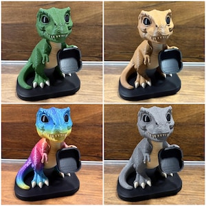 May include: A set of four dinosaur-shaped stands, each featuring a textured body, large black eyes, and a small black square tray held in the front. The figures are available in solid green, tan, gray, and a multi-colored gradient finish on a black base.