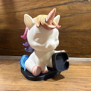 Puede incluir: Una figura de unicornio blanca con cuerno y pelo de color cobre metálico, con una crin degradada que va del rosa al morado y al azul. El unicornio está sentado sobre una base negra y sostiene un soporte cuadrado negro con una ranura.