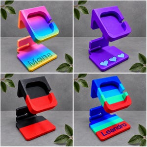 Op de afbeelding: Vier Z-vormige oplaadstandaards voor smartwatches. Eén is regenboogkleurig met de naam Mona, één is effen paars met drie hartjes, één is zwart met rood, en één is blauw, groen en rood met de naam Leandro op de basis gedrukt.