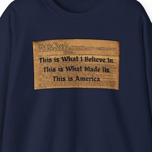 May include: Navy blue sweatshirt with a rectangular graphic featuring the text "We the People" and the phrases "This is What I Believe In," "This is What Made Us," and "This is America" in black font. The graphic has a parchment paper appearance.