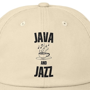 以下が含まれることがあります： ベージュのダメージ加工が施されたベースボールキャップに、黒い文字で「JAVA AND JAZZ」と書いてあります。テキストの上に、音楽記号がついたスタイリッシュなコーヒーカップがあります。