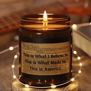 May include: A brown glass candle with a white label that reads "We the People" and "This is What I Believe in. This is What Made Us. This is America." The candle is lit and sitting on a wooden coaster.