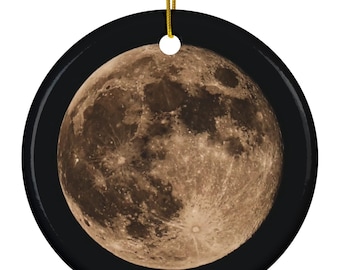 May include: A round ceramic ornament featuring a detailed depiction of the moon against a black background. The moon is rendered in shades of brown and tan, with visible craters and textures. A gold string is attached for hanging.