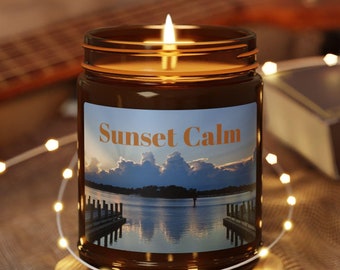Puede incluir: Una vela de vidrio marrón con una etiqueta que dice "Sunset Calm". La etiqueta presenta una fotografía de un lago con un muelle y un cielo de puesta de sol. La vela está sentada sobre un posavasos de madera y rodeada de luces blancas.