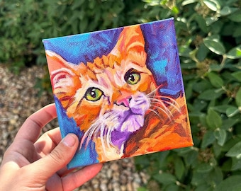 以下が含まれることがあります： 緑色の目をしたオレンジ色のトラ猫の小さな絵画。猫は青、紫、オレンジの背景のキャンバスに描かれています。