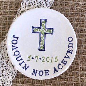 May include: A white ceramic plate with a blue and green cross design. The cross is filled with a green leaf pattern and outlined in blue. The plate has the name "JOAQUIN ACEVEDO" and the date "5.7.2016" in blue and green lettering.
