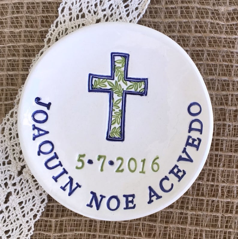 May include: A white ceramic plate with a blue and green cross design. The cross is filled with a green leaf pattern and outlined in blue. The plate has the name "JOAQUIN ACEVEDO" and the date "5.7.2016" in blue and green lettering.