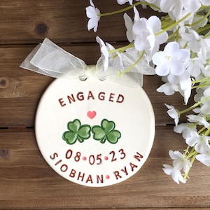 May include: A cream-colored ceramic engagement ornament with the word "ENGAGED" in brown lettering. It features two green shamrocks, a pink heart, the date "08-05-23", and the names "SIOBHAN RYAN". A sheer ribbon is tied at the top.
