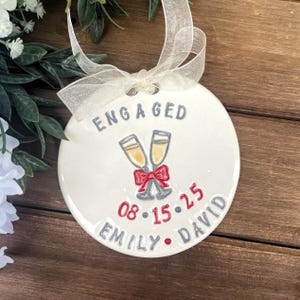 May include: White ceramic engagement ornament with the word "ENGAGED" in gray. Features two champagne glasses with a red bow, date "08.15.25", and names "EMILY • DAVID". Includes a sheer ribbon for hanging.