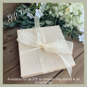 Puede incluir: Una caja de regalo cuadrada, de color marr&oacute;n claro, atada con una cinta transparente de color crema. La caja est&aacute; sobre una superficie de madera, con flores blancas y follaje verde en el fondo. El texto en la caja dice "Gift Box".