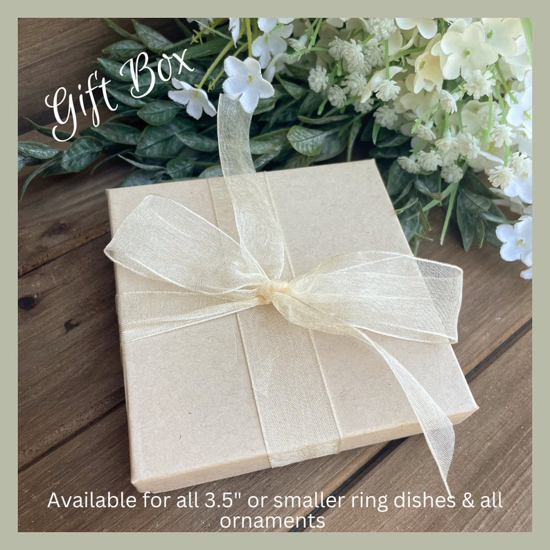 Puede incluir: Una caja de regalo cuadrada, de color marr&oacute;n claro, atada con una cinta transparente de color crema. La caja est&aacute; sobre una superficie de madera, con flores blancas y follaje verde en el fondo. El texto en la caja dice "Gift Box".
