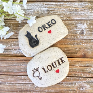 May include: Two speckled, light-colored stones with black text and cat illustrations. One stone reads "OREO" with a black cat and red heart. The other reads "LOUIE" with a cat-shaped design and red heart. White flowers are in the background.