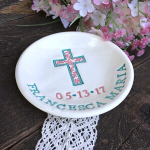 May include: A white ceramic dish with a teal cross design filled with pink floral patterns. The dish has the name "FRANCESCA MARIA" and the date "05-13-17" in teal and pink lettering. Decorative floral accents are in the background.